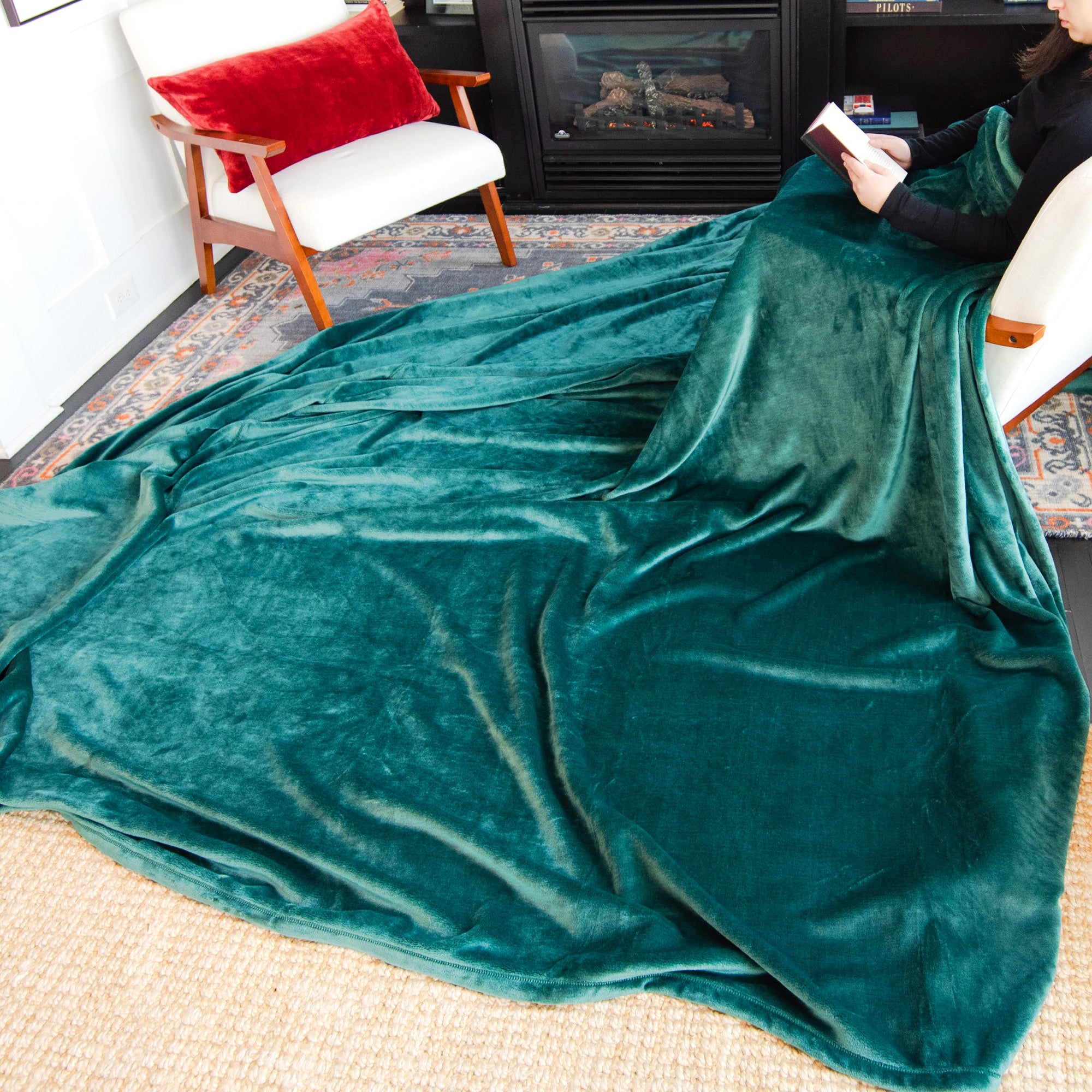 American Blanket Company - Luster Loft Oversized Fleece Blankets - Pictured young woman reading in front of a fireplace and have evergreen luster loft fleece blanket cascading over her lap and over the floor by 2-3 feet