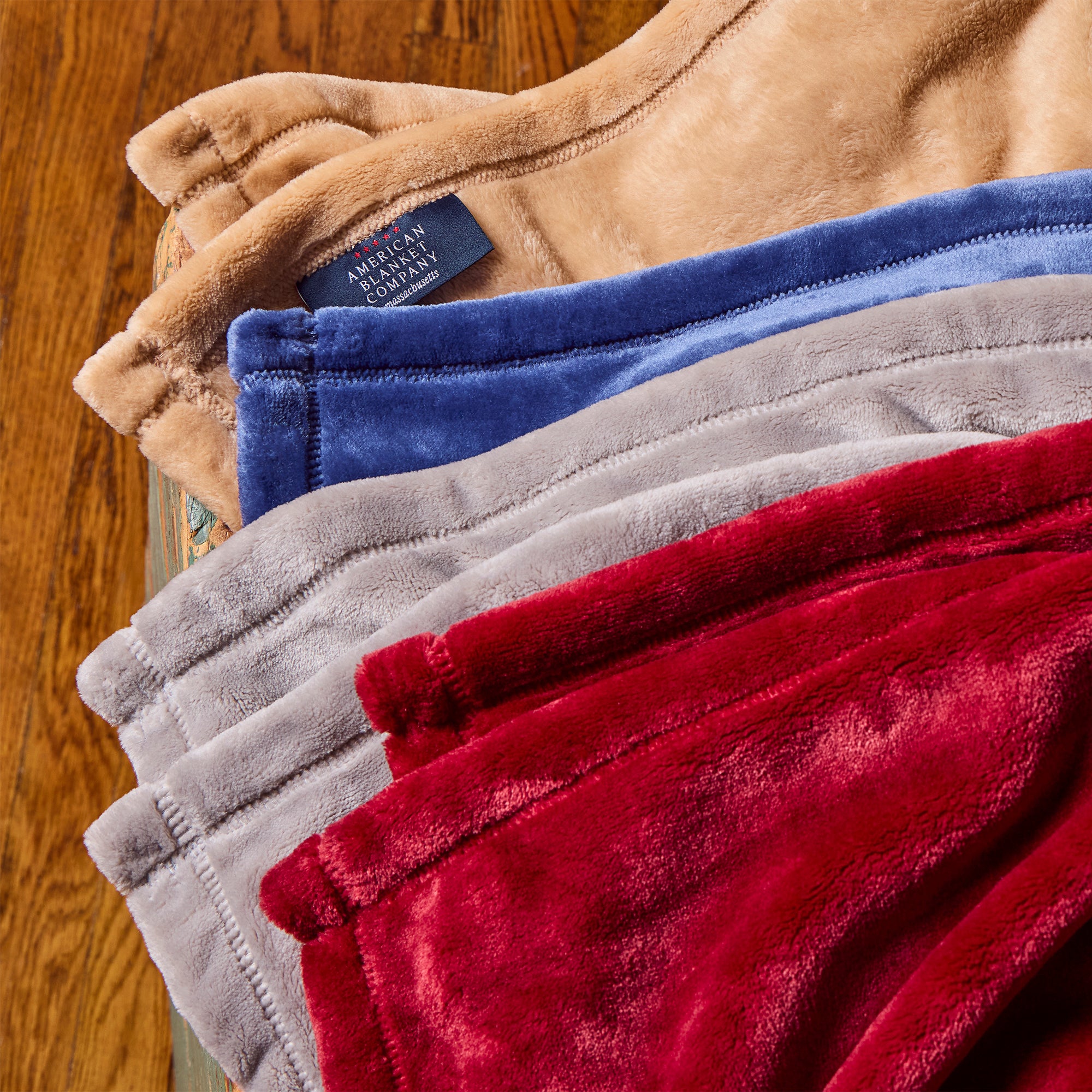American Blanket Company - Luster Loft Fleece Oversized Fleece Blankets - Stack of folded fleece blankets in various colors with a brand label on a wooden surface.