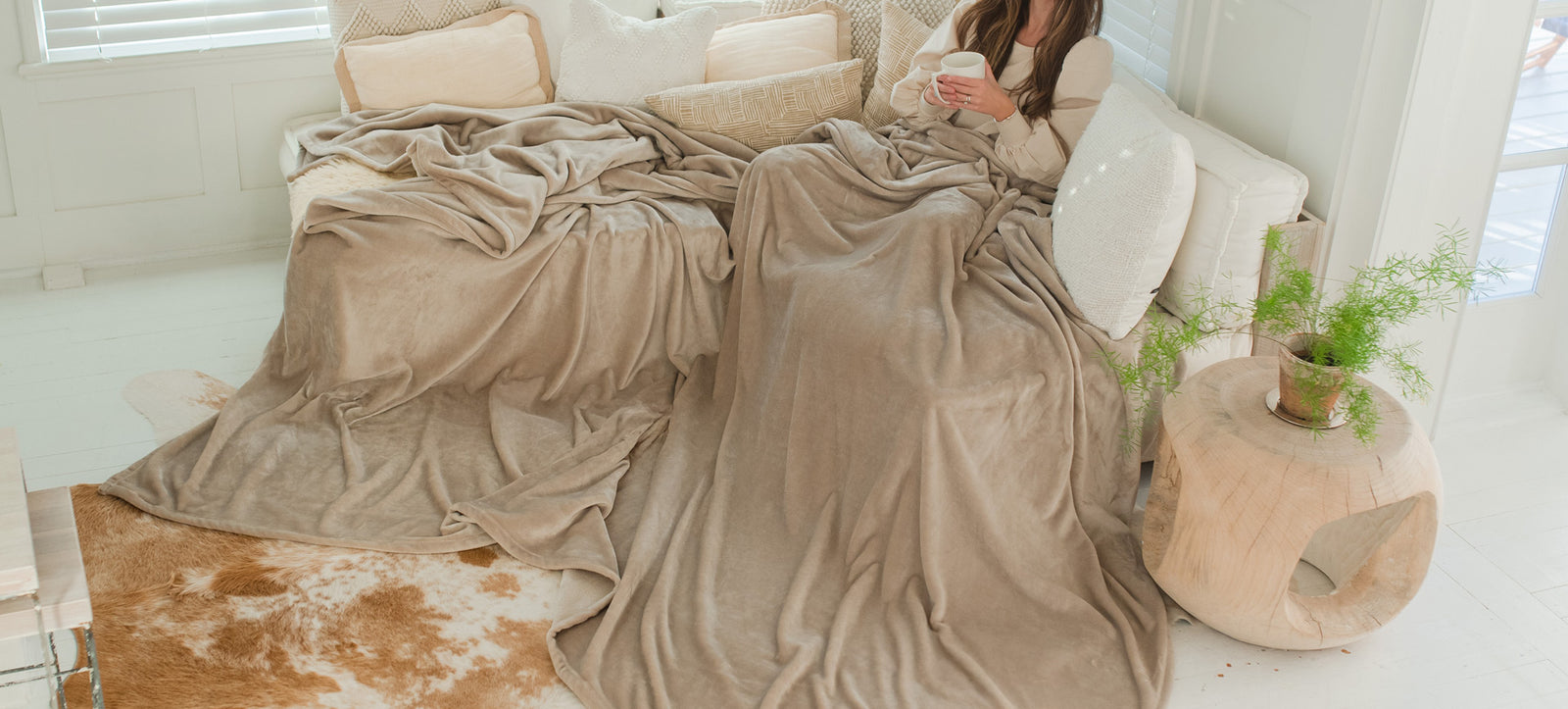 AMERICAN BLANKET COMPANY - A WOMAN HOLDING A MUG ON A COUCH, BUNDLED UP WITH A LUSTER LOFT OVERSIZED FLEECE BLANKET IN LATTE