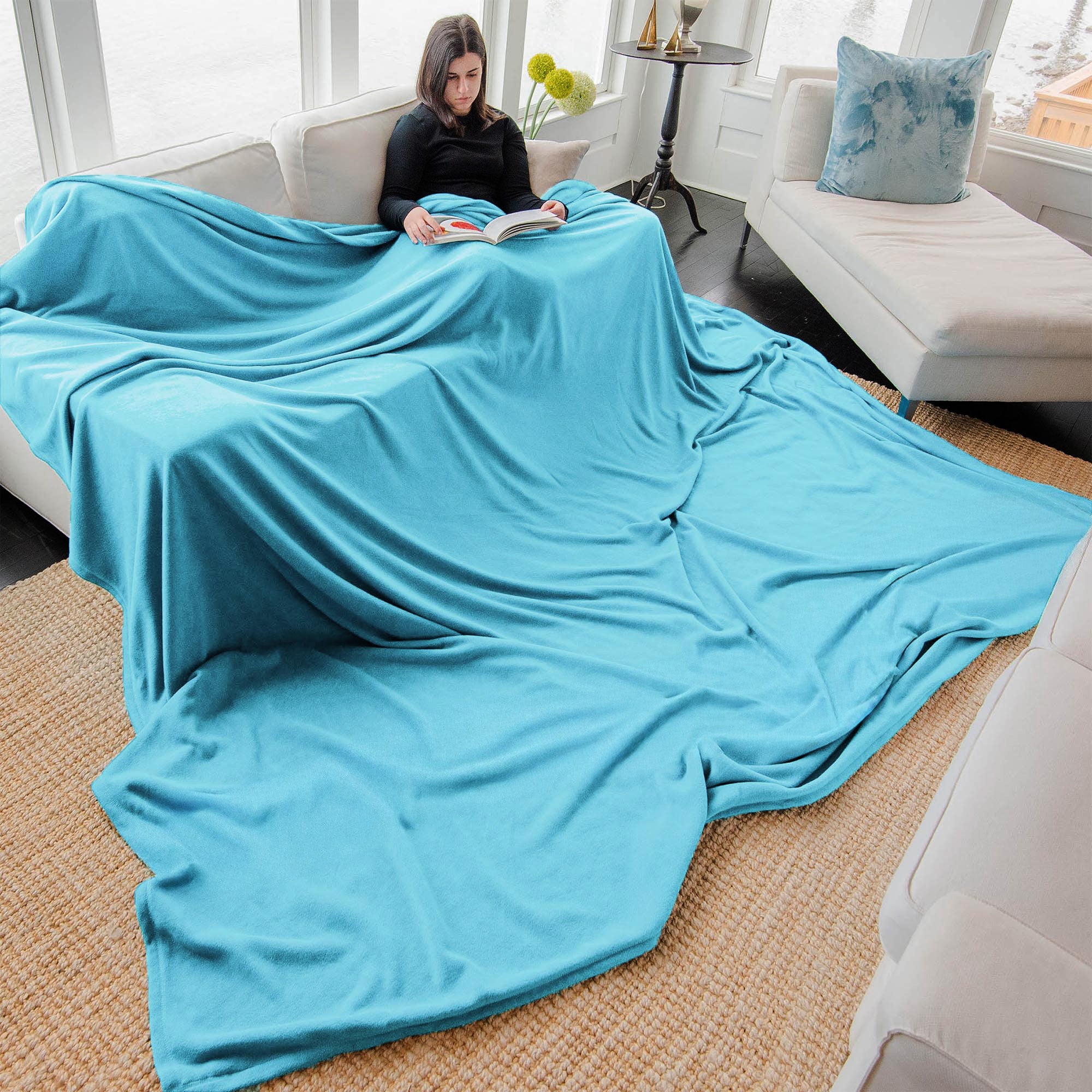 American Blanket Company - Peaceful Touch Fleece Blankets - Oversized Custom - Pictured is a young woman reading on a couch in a living room with a large peaceful touch oversized blanket in light blue.