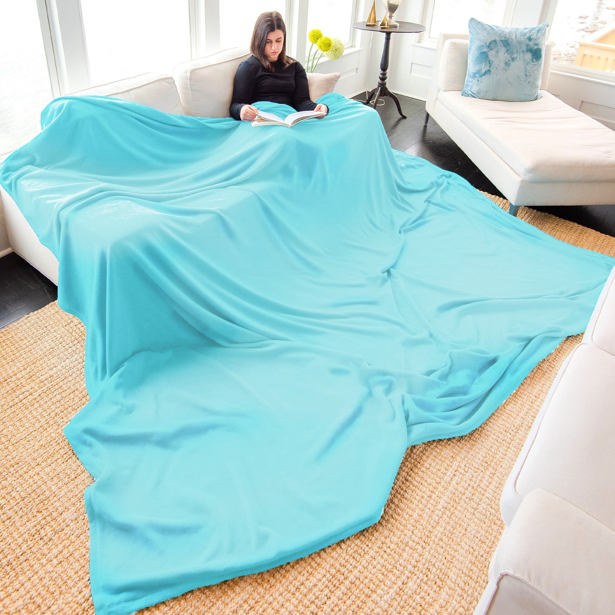 American Blanket Company - Peaceful Touch Biggest, Oversized, Fleece Blankets - Young woman reading on a couch with an oversized peaceful touch light blue blanket flowing onto the floor.