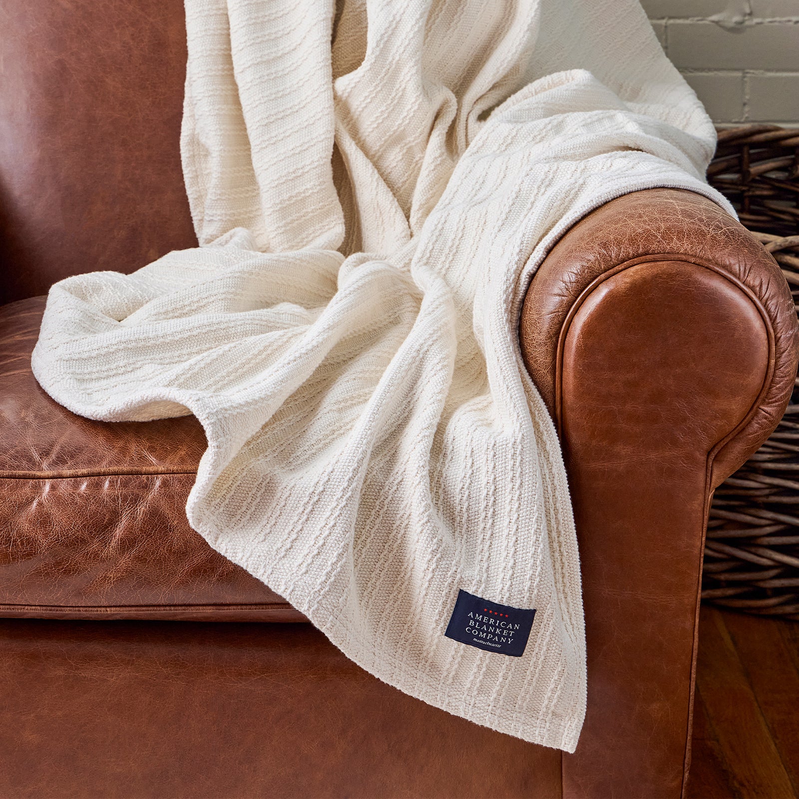 American Blanket Company - Oversized Cable weave 100% Cotton Blanket -  Cable Weave Cotton Blanket - White textured blanket draped over a brown leather armchair with a visible brand label.