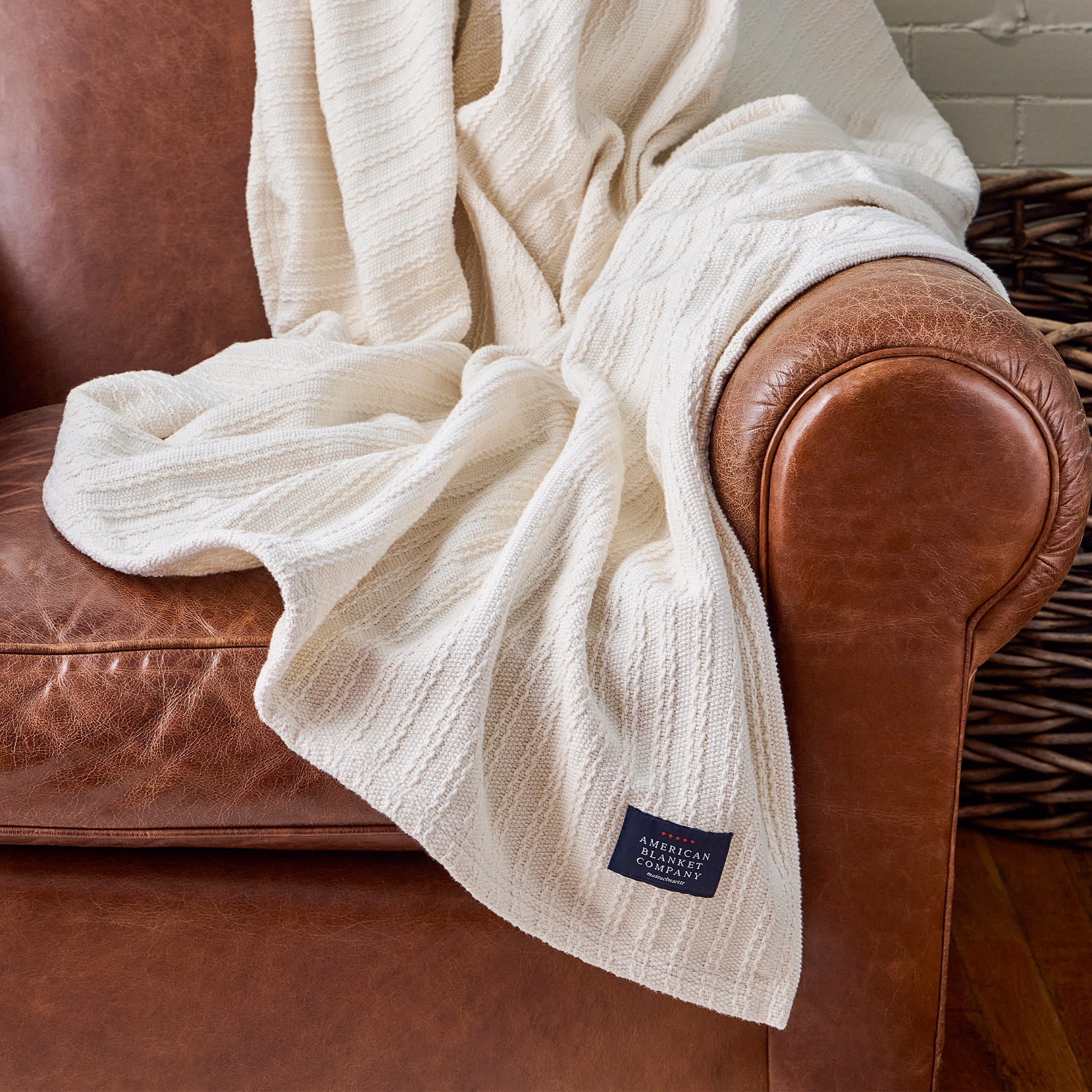 American Blanket Company - Oversized Cable weave 100% Cotton Blanket -  Cable Weave Cotton Blanket - White textured blanket draped over a brown leather armchair with a visible brand label.