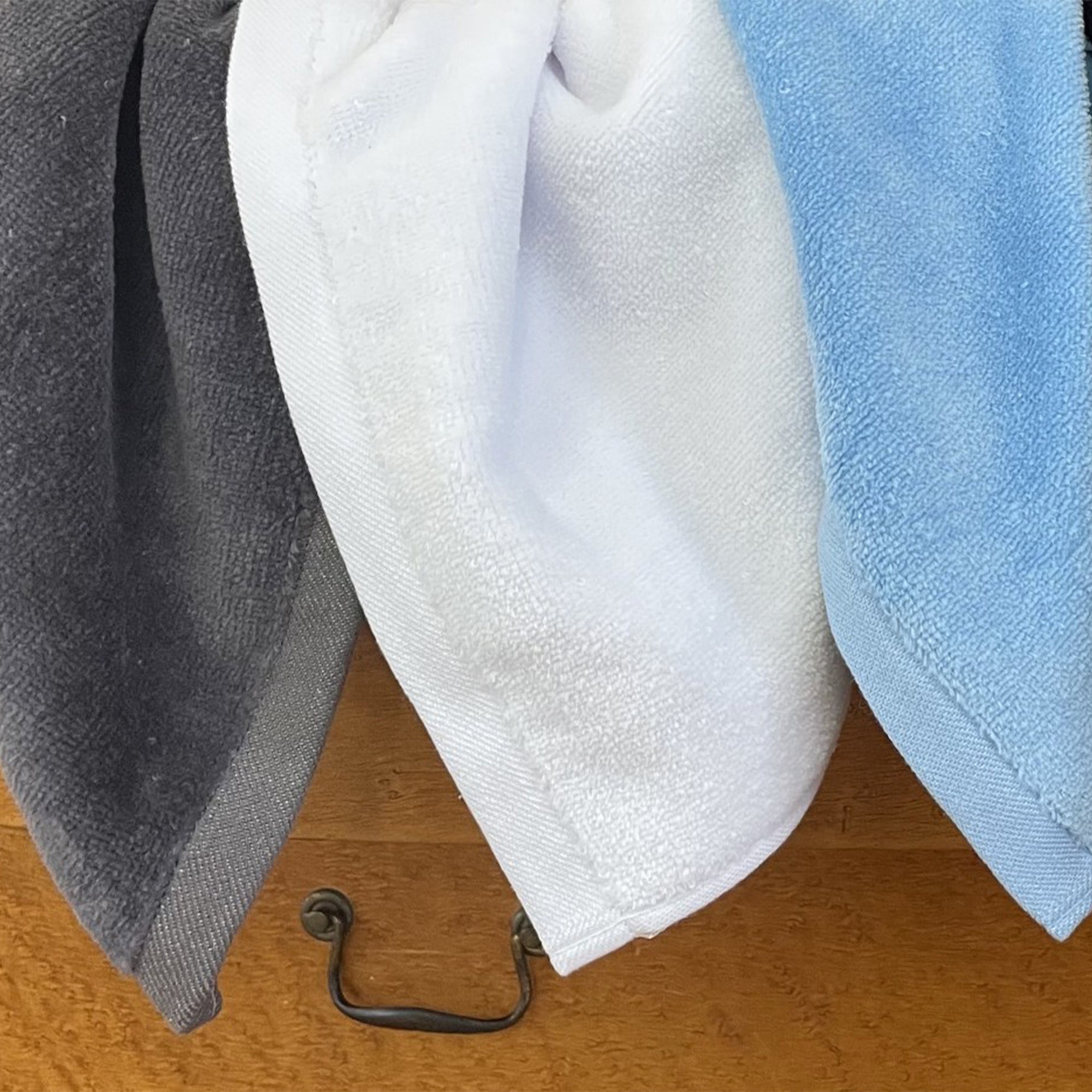 American Blanket Company -  Colorful Made in the USA 100% Cotton towels - Three towels in gray, white, and blue hanging on a wooden surface with a metal hook.