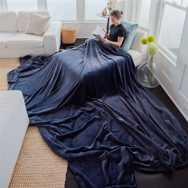 American Blanket Company - Luster Loft Fleece Oversized Fleece Blankets - Woman sitting on a  chaise lounge with a large blue fleece blanket spilling out onto the floor.