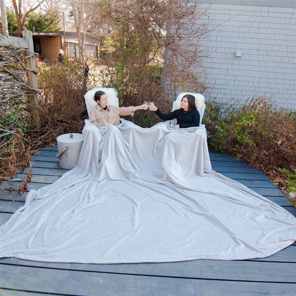 American Blanket Company - Luster Loft Oversized Fleece Blankets - Pictured are two woman toasting outside on a blue wooden deck, both covered by a large luster loft blanket cascading over both of the laps over the blue deck.