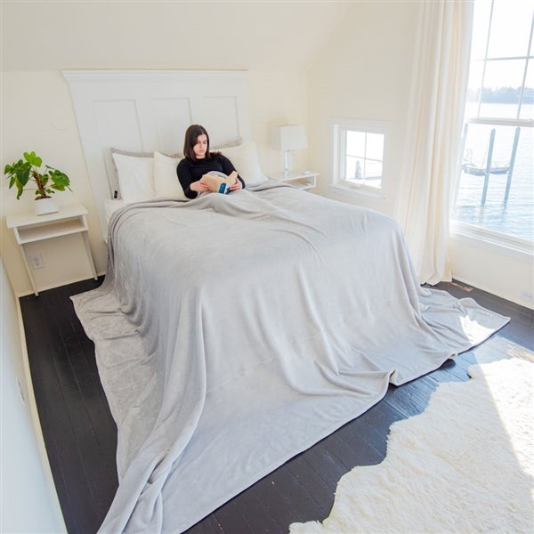 American Blanket Company - Luster Loft Oversized Fleece Blankets - pictured young woman on a large bed draped in a light gray luster loft oversized blanket that falls over the bed and past it onto the floor.