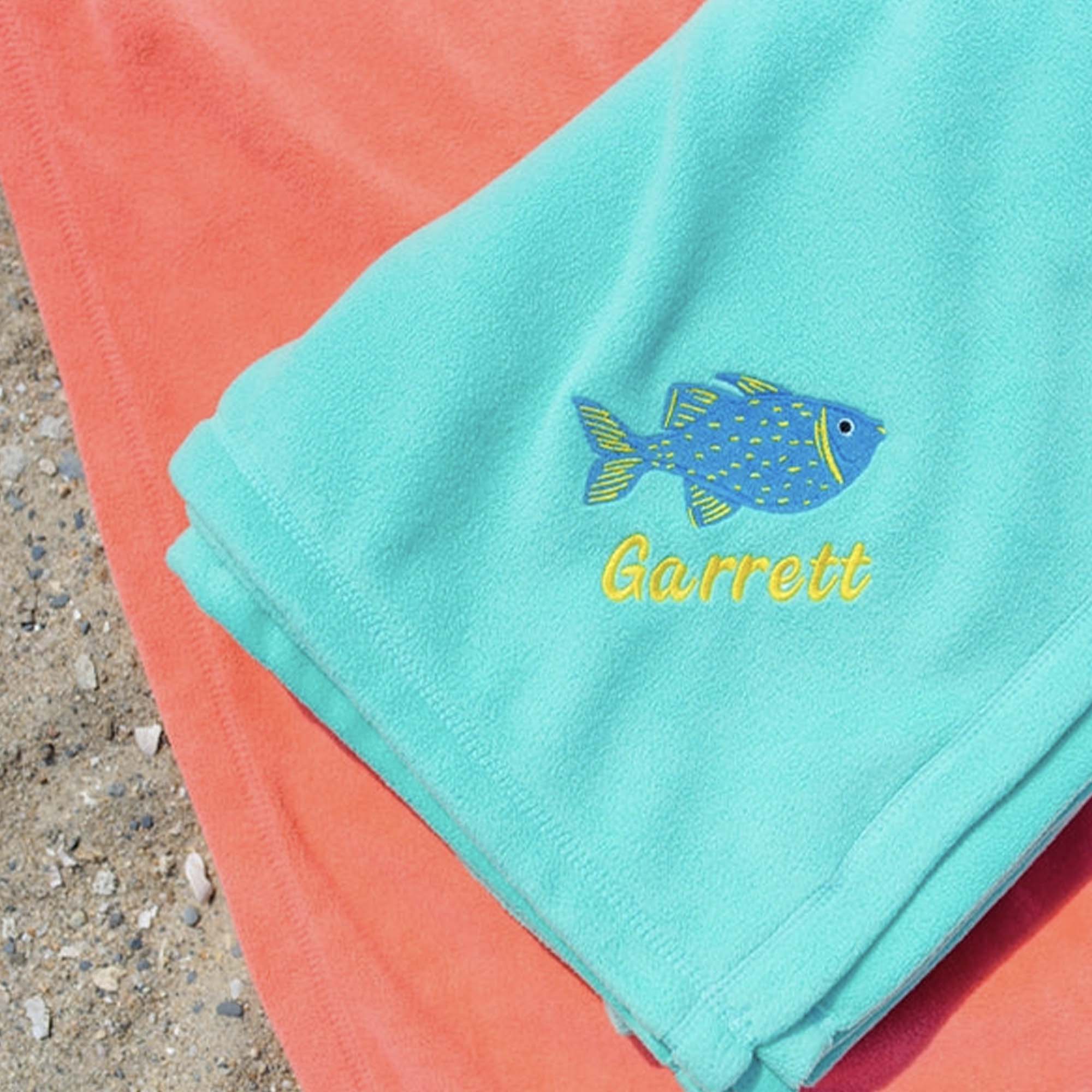 American Blanket Company - Peaceful Touch Beach Blankets - Pictured is a pair of peaceful touch beach blankets, coral and light blue with a blue fish embroidery and a name under it.